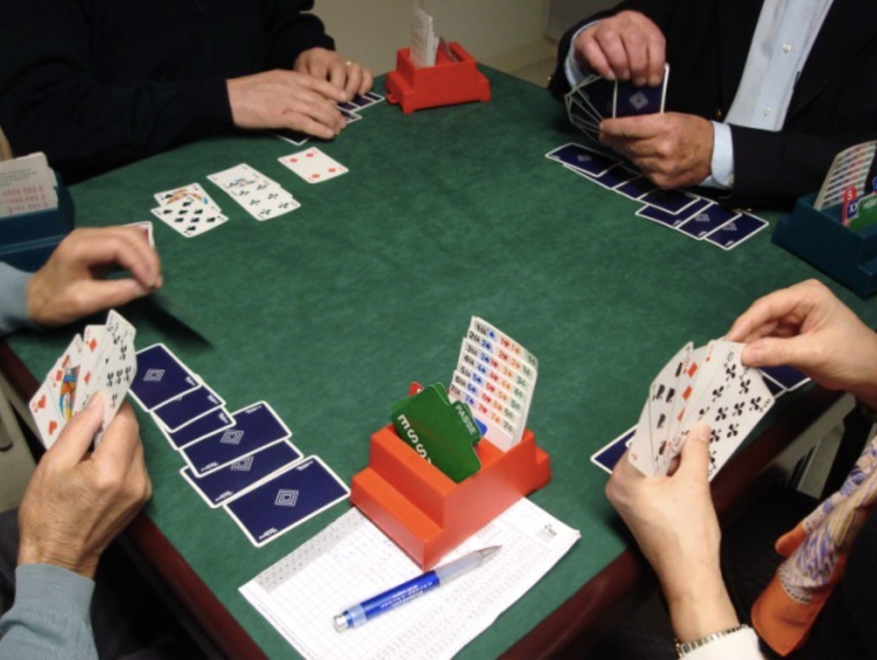 Quatre personnes autour d'une table pour jouer au bridge avec des cartes, des étuis et des boites d'enchères.
